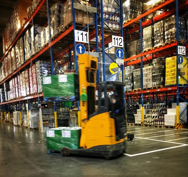 Forklift carrying a wrapped pallet between tall racks of stored goods in a warehouse